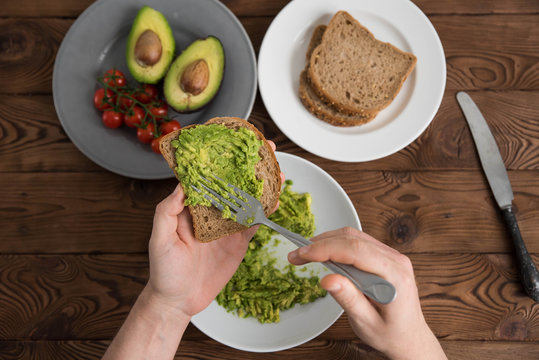 Hands of cook chef preparing healthy avocado sandwich on dark rye toast bread made with fresh avocado paste, cherry tomatoes on brown wooden background - Powered by Adobe
