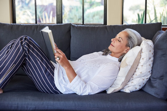 Mature woman with grey hair reading a book lying down on sofa in living room