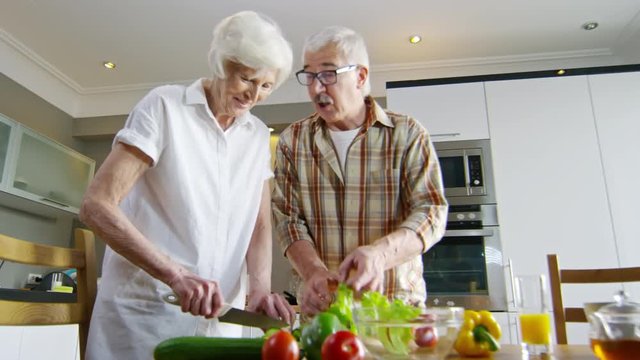 Low angle of old woman and man standing at kitchen table and cutting vegetable salad together and talking