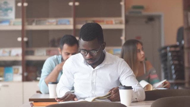 education, university and people concept - attentive african american adult man student reading book sitting at the desk at library with other people on background