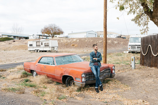 Young adult male in denim sits in front of broken down vintage car in rural town