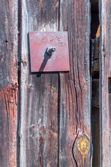 A close up of a metal anchor rod is mounted to the outside of a rustic old barn by a nut and square red and rusted mounting plate. The siding of the barn is aged and textured.