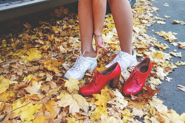 Young woman putting on white sports shoes for active walking in park. Legs of single girl in run snickers on the road among falling yellow maple leaves. Red woman boots wiht high heels. Autumn day.