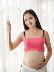 Asian pregnant woman touching her belly near window, lifestyle concept.