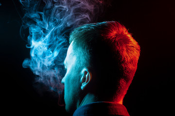 A view from the back of a boy’s head around in a shirt smoking a vape and exhaling multi-colored...