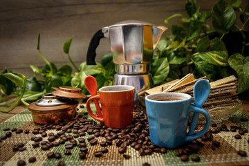 Orange and blue cups of coffee, moka pot coffee maker, old alcohol stove and cookies. Wooden table with bamboo mat and coffee beans spilled on the table.