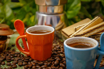 Orange and blue cups of coffee, moka pot coffee maker, old alcohol stove and cookies. Wooden table with bamboo mat and coffee beans spilled on the table.