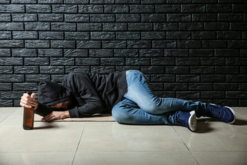 Depressed young man with bottle of beer lying on floor near black brick wall. Alcoholism concept
