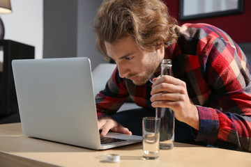 Young man using laptop and drinking vodka at home. Alcoholism concept