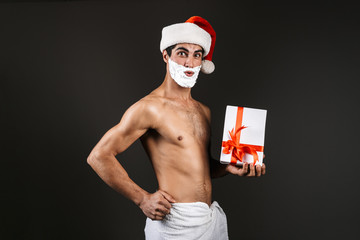 Young santa naked man posing isolated over dark wall background holding gift box dressed in towel with shaving gel on beard.