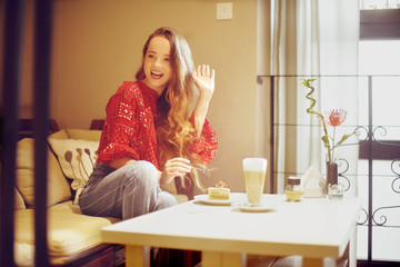 The girl in the bakery eats dessert. Beautiful model in a cafe eats sweets and smiling. Beautiful girl in a red sweater with long hair eats in a restaurant alone, diet. Girl drinking coffee /latte