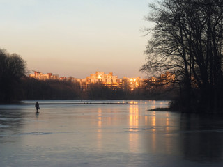 Fototapeta premium A woman crosses a city pond on thin ice at sunset on the background of orange facades of a residential area.