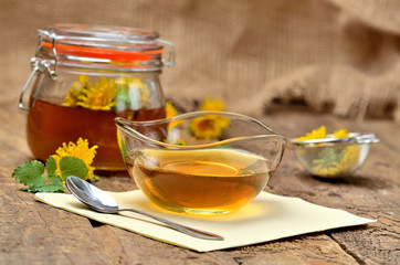 Fototapeta na wymiar Dandelion jam in glass, spoon, dandelion head around, small colander and full jar of jam in background