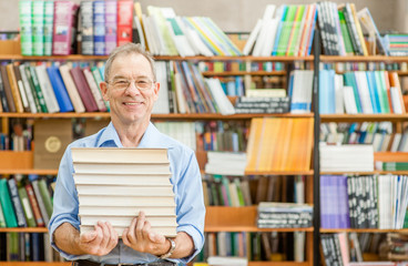 Happy senior man holds a pile of books in the library. Empty space for text