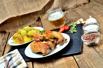 Golden grilled chicken legs on a plate, roasted potatoes and cold beer, rosemary, tomatoes, oregano, salt, pepper and garlic on a wooden table