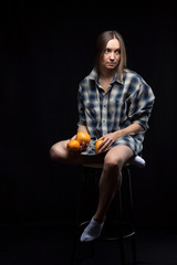 A girl in a shirt is sitting on a chair holding oranges in her hands on a black background.