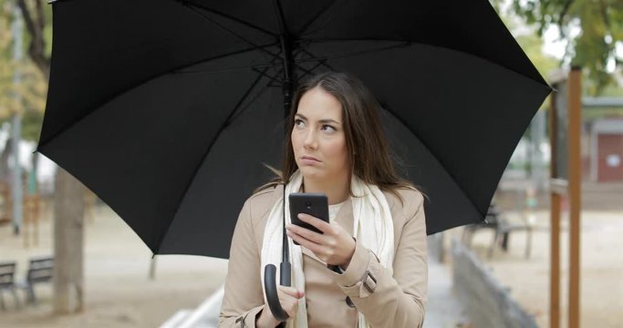 Front view portrait of a frustrated woman checking weather app on a smart phone under the rain in winter