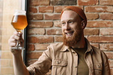 Portrait of young bearded hipster man with happy emotional face holding glass with craft beer,...