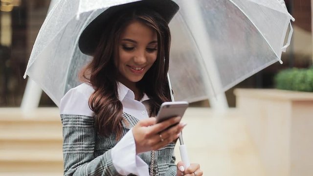Beautiful young business woman using smartphone on the street in rainy weather, smiling, holding umbrella, communication concept