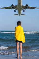 Back view on the boy in the yellow towel standing on seashore under landing plane. Travel Concept.