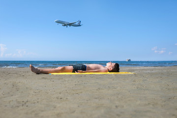 Fototapeta na wymiar Teen boy lies on yellow towel and sunbathes on the beach under the landing planes. Traveling on an airplane with children. Concept