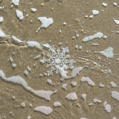 Fototapeta na wymiar Huge sparkling snowflake on the sand in the sea foam. Concept of Winter and Christmas vacation on the beach and resort.