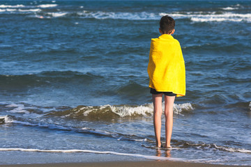 Fototapeta na wymiar Back view on the boy in the yellow towel standing on seashore. Travel Concept.