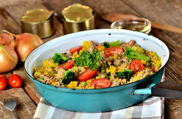 Tuna risotto with vegetables, tomatoes, broccoli and parsley in the old pan, onions, cans and oil in the background