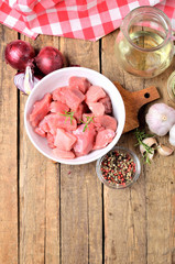 Top view of raw pork meal diced in a bowl, garlic, half of onion, pepper, jug with oil and checkered red tablecloth in the background - vertical photo