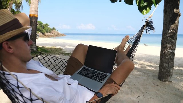 Young Business Man With Laptop Swinging In Hammock On The Beach