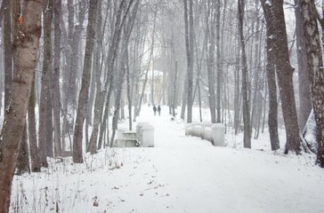 a winter park with a bridge and a road to the manor, two people are walking along the road, the trees are covered with snow, snow is falling.