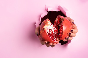 Woman hands holding ripe pomegranate fruit through torn pink paper background. Fresh fruit juice....