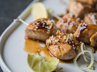 Grilled Asian teriyaki chicken skewers with sesame, leek and citrus juice on a white oval plate on dark grey background