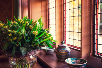 Close up bouquet of fresh yellow flowers in glass vase near a wooden window with ceramic medieval style dishes on the windowsill. Elegant comfort concept. Selective focus. Copy space.