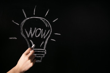 closeup female hand drawing a white chalk light bulb on a black board with the word wow