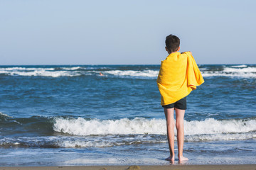 Fototapeta na wymiar Back view on the boy standing on seashore of the beach in the yellow towel and looking on the sea. Concept.
