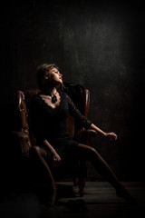 Fototapeta na wymiar Roaring Twenties. Woman portrait in the style of Gatsby. Low key. A beautiful young woman in a black lace dress is sensually sitting in a brown leather chair.