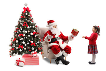 Santa Claus sitting in an armchair next to a Christmas tree and giving a present to a surprised little schoolgirl