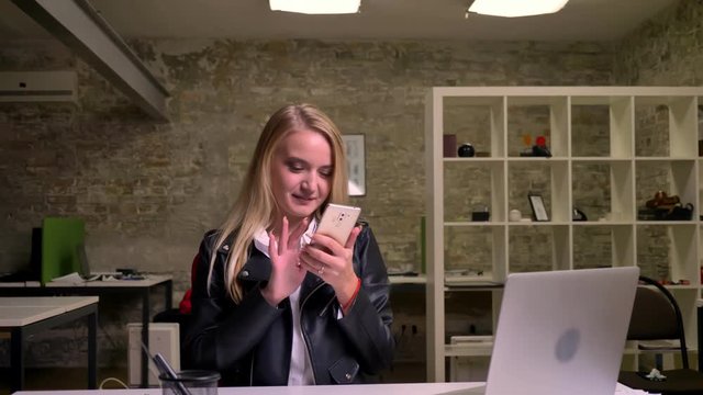 Happy smiling caucasian blonde girl is sittinf at desktop at taking selfies on her smartphone while hanging in light brick office