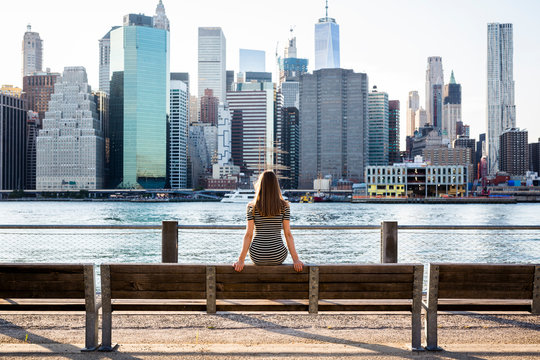 USA, New York, Brooklyn, back view of woman sitting on bench in front of East River and skyline of Manhattan