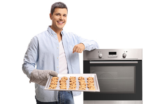 Young man holding a tray of freshly baked cookies and leaning on an oven