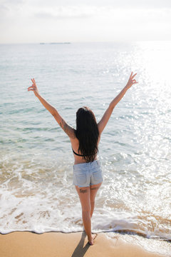 Back view of young woman standing at seashore showing victory sign