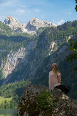 Naklejka na ściany i meble Young beautiful woman sitting on a rock looking out at a mountain waterfall over a reflecting mountain lake