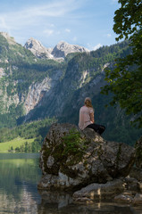 Young beautiful woman sitting on a rock looking out at a mountain waterfall over a reflecting mountain lake