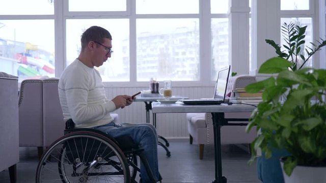 online learning, freelancer man disabled in wheelchair wearing glasses uses a cell phone sitting at a table with computer laptop in a cafe