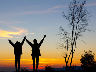 Silhouette of happy two girls standing with raised hands on the mountain at the sunset or sunrise time.