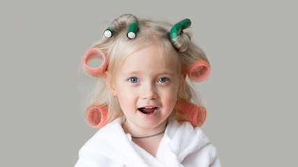 Happy little caucasian girl with hair curlers. Beautiful blonde girl with hairpin in her hair with a white robe after a bath.
