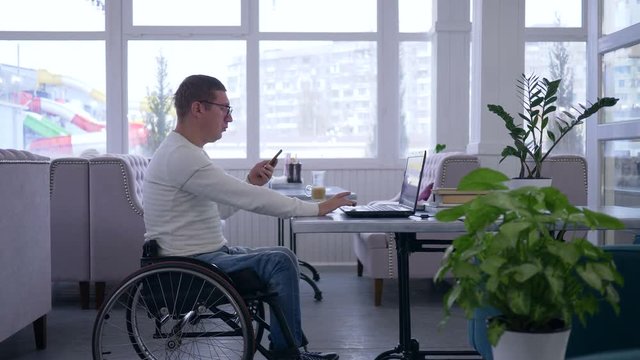 remote business management, handicapped man in wheelchair uses a cellphone sitting at a table with laptop in a cafe on the background of a large window