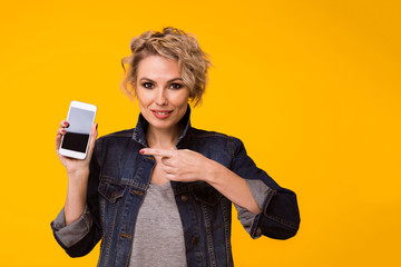 happy blond woman in sweater showing blank smartphone screen and pointing on it while looking at...