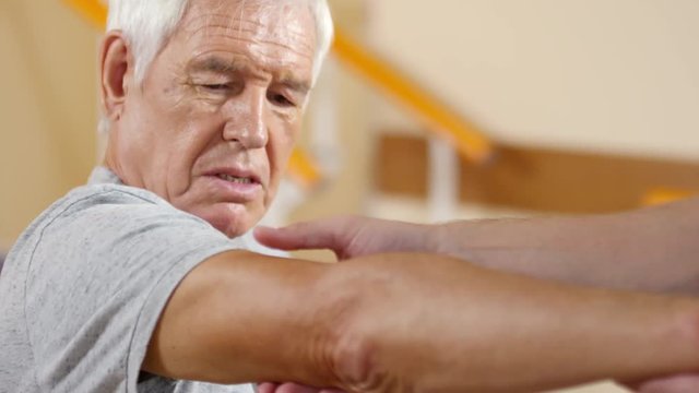 Medium shot of senior man with grey hair talking to unrecognizable male physical therapy specialist examining his arm
